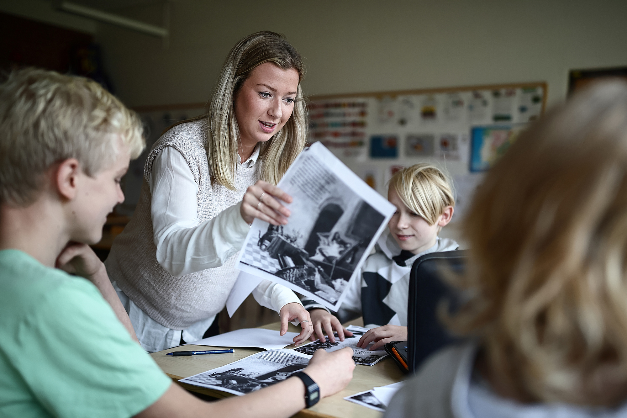 En lärarstudent delar ut papper till 3 elever som sitter och arbetar vid ett bort. 